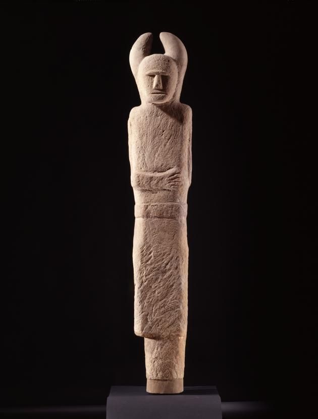 Tall stone statue of a stylized human figure with a cylindrical body, abstract facial features, and two upward-curving elements above the head.
