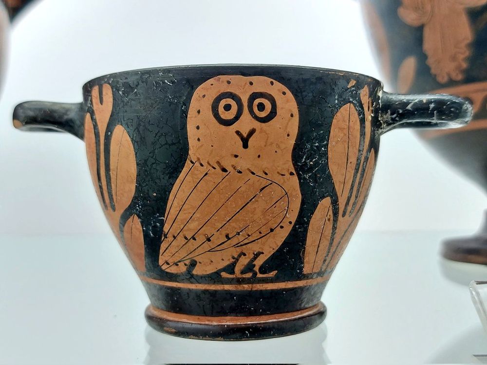 A red-figure cup with two handles, featuring a central depiction of a stylized owl flanked by olive leaves. The owl is painted in a reddish-brown hue against the black background. 