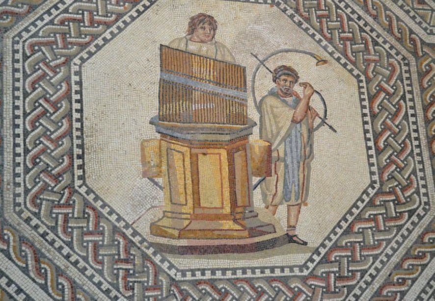 A Roman mosaic featuring two figures. One person is playing a  hydraulis (water organ), while the other stands beside it holding a curved horn. The mosaic is framed by a decorative geometric border with interwoven patterns in red, black, and white.