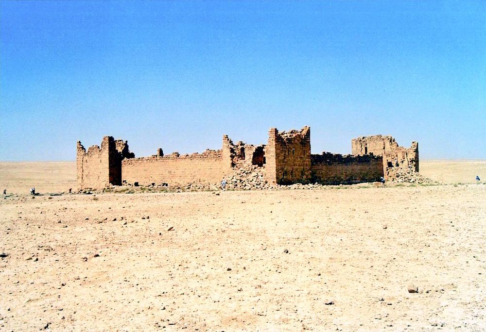 Ruins of an rectangular desert fort with partially collapsed towers, surrounded by empty, rocky sand and no vegetation.