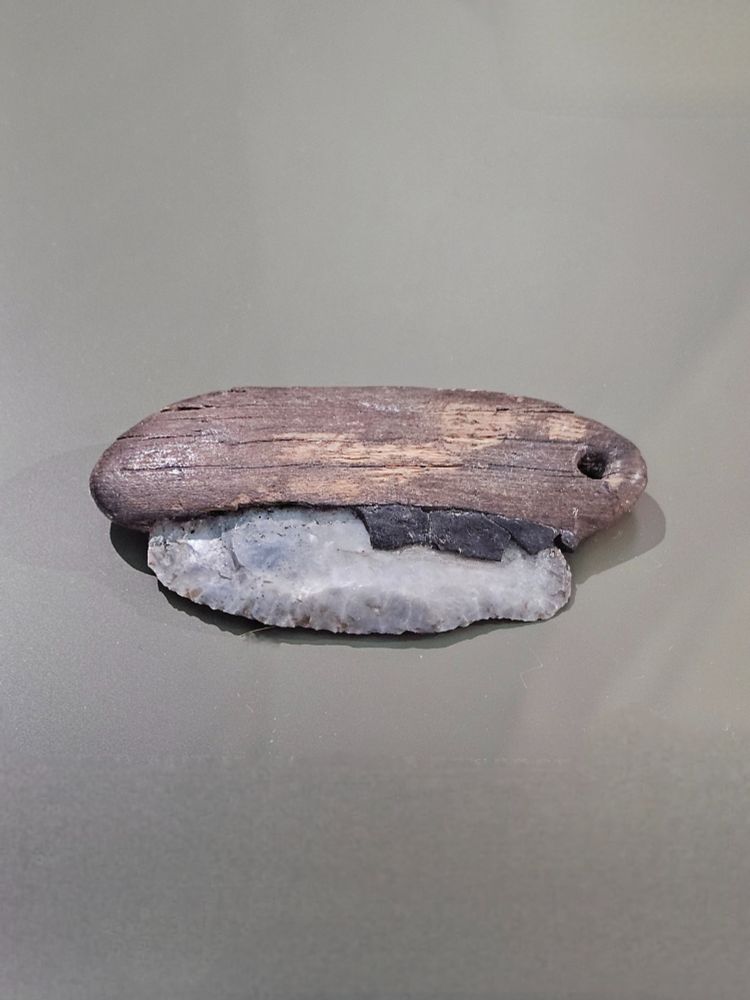 A flint knife featuring a curved stone blade secured into a wooden handle using black birch tar adhesive. The dark handle has a small perforation at one end. The tool is displayed on a neutral surface, showing the craftsmanship and natural materials used.