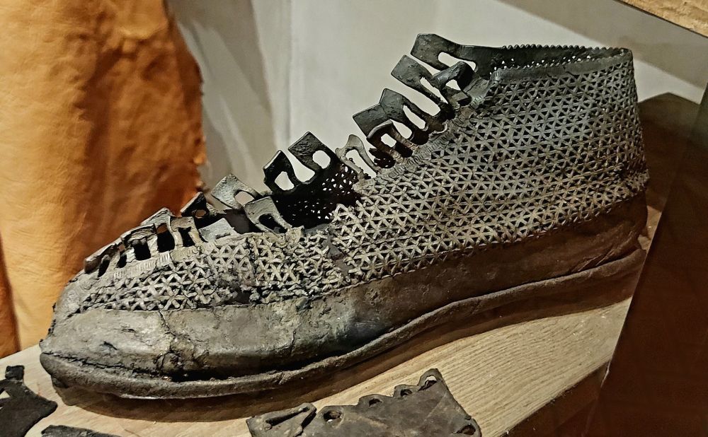 A well-preserved Roman leather shoe displayed in a museum setting. The shoe features an intricate lattice-like pattern with small, repeated triangular cutouts across the upper section. The toe is rounded and the sole appears worn and cracked with age. The top has a series of slits likely used for lacing. The leather is darkened from time and exposure, giving it a textured, weathered appearance.
