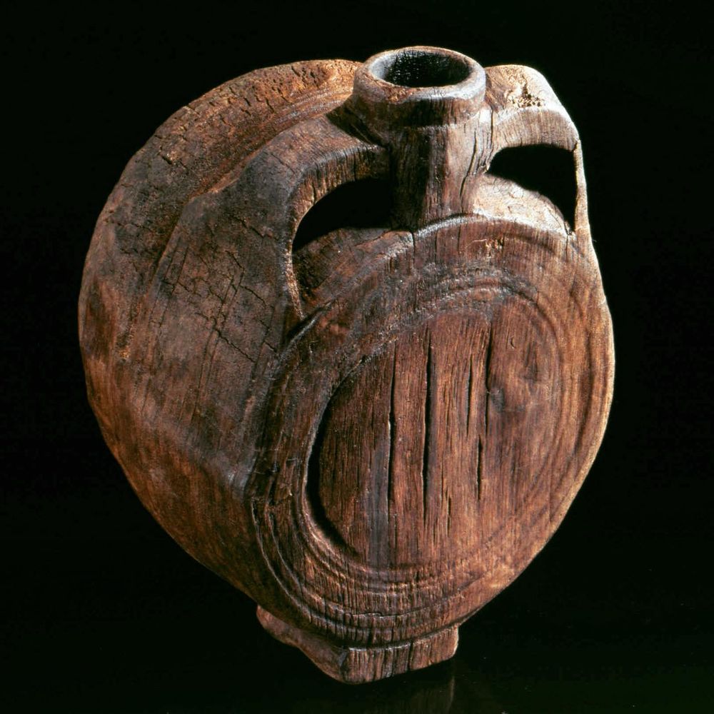 A weathered, round wooden canteen with a flat base, central spout, and two carved side handles. The surface shows deep cracks and a rough texture, indicating age and use. The canteen has a barrel-like shape, photographed against a black background.