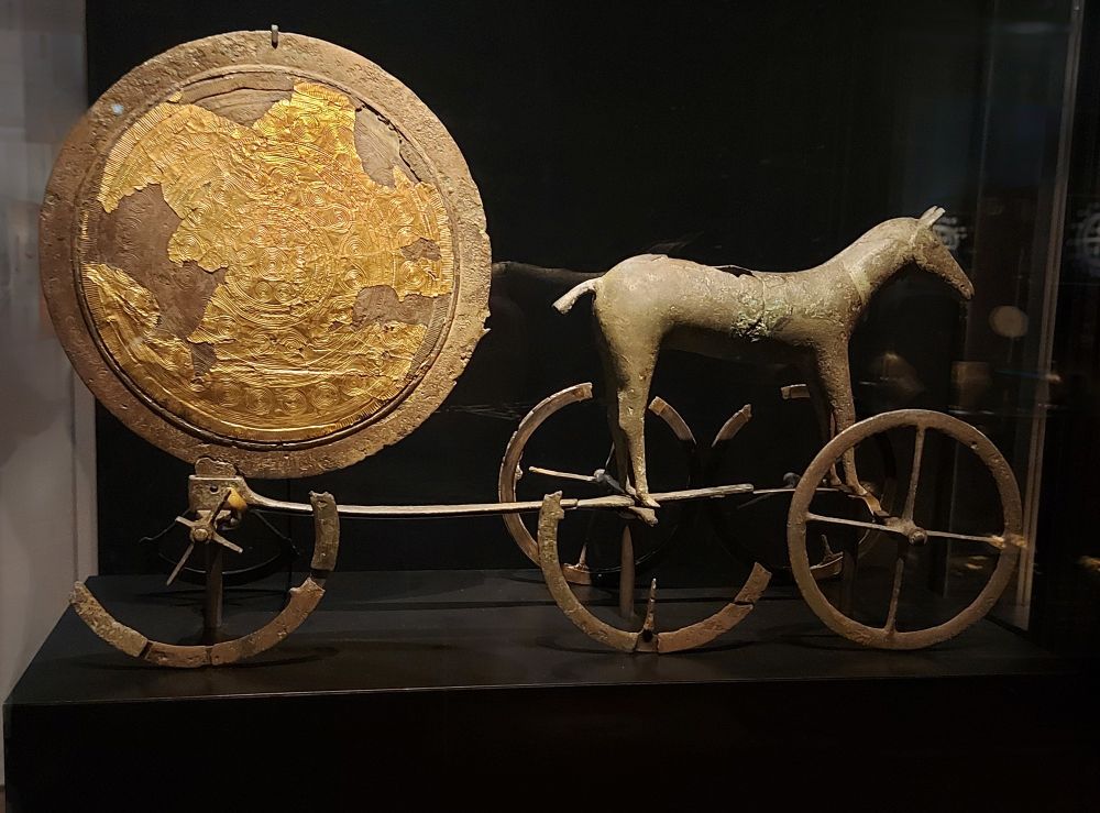 Photo of the Trundholm Sun Chariot, featuring a horse figure pulling a large, gilded bronze disk on a wheeled frame. The disk is partially covered in gold leaf with intricate circular patterns, symbolizing the sun. The sculpture is mounted on six spoked wheels and displayed against a dark background.