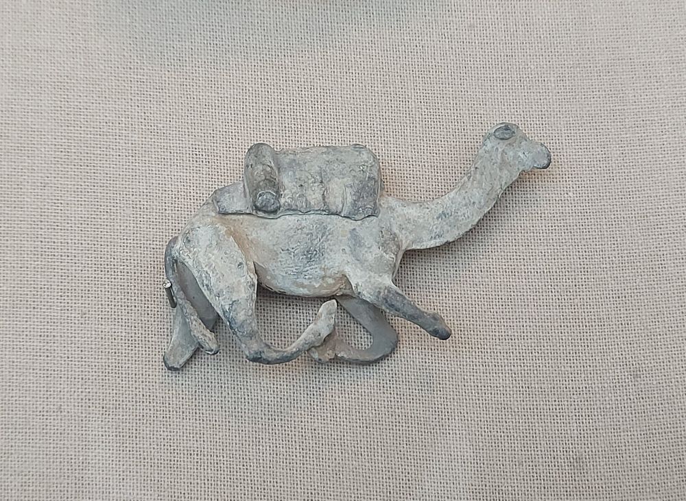 An lead figurine of a camel carrying a load on its back, displayed against a beige fabric background. The camel is depicted in motion, with its legs bent and extended, and has a long, curved neck with a slightly raised head. The load on the camel’s back is rectangular with a cylindrical object on top, possibly representing a pack or rolled goods.