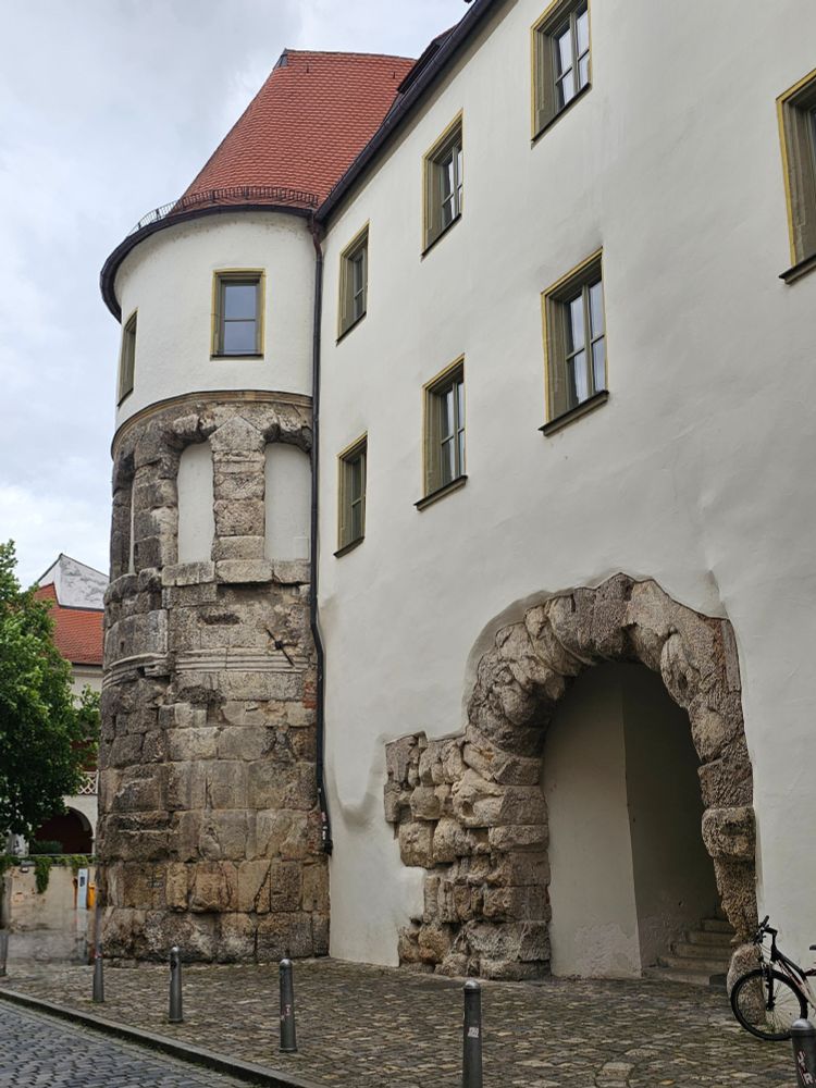 A photo of the gate tower and the gate way integrated in the former Bishop's residence: The lower section features rough stone masonry and a partially preserved archway, while the upper structure has smooth white walls, rectangular windows with yellow trim, and a red-tiled roof. The historic elements are visibly integrated into the facade along a cobblestone street.