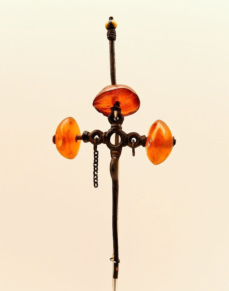 A decorative bronze pin featuring a vertical rod with a central horizontal bar adorned with three large, translucent amber-colored beads. A small chain hangs from the center, and the top is capped with a dark finial and bead-like ornament.