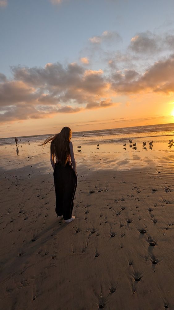 A teenager with long brown hair and baggy sweatpants stands on a beach at sunset
