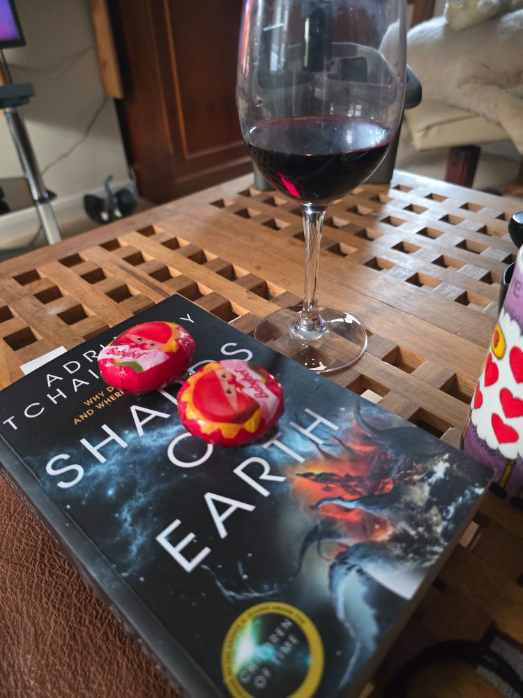 Two baby bell cheeses rest on a book. The book is a soft back copy of Shards of Earth by Adrian Tchaikovksy. A glass of red wine sits on the table next to the book.