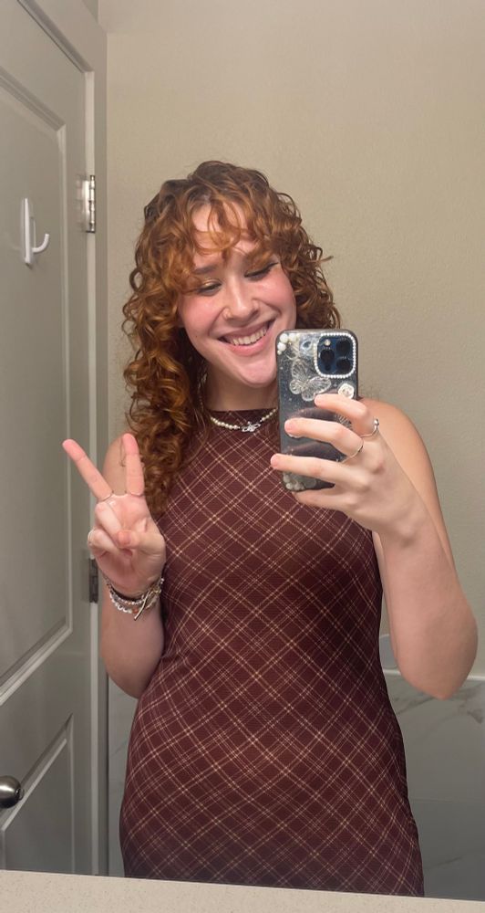a mirror selfie of capri holding up a peace sign and smiling. she’s wearing a brown plaid dress