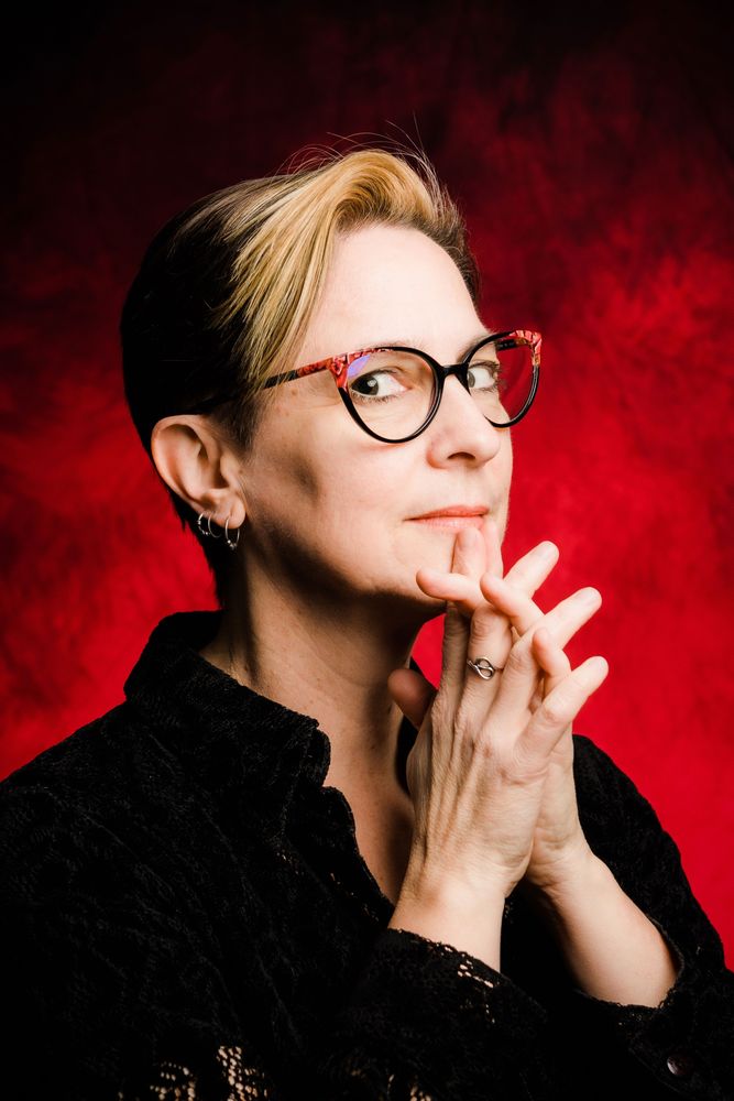 Vicki Walden has short blonde hair, wears a black shirt and red & black glasses. Her hands are clasped together under her chin, fingers steepled, while she looks at the camera.