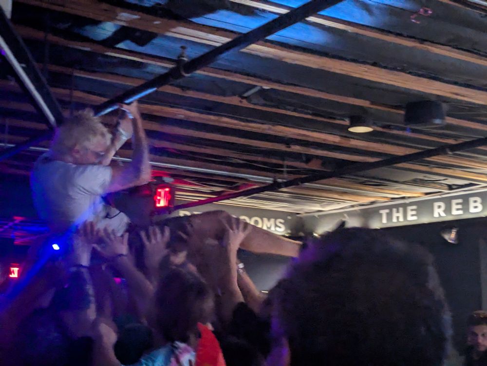 Picture of Frankie crowd surfing at his bands show. He is holding on to a pipe for the sprinkler system. You can see a crowd of people desperate to touch him 