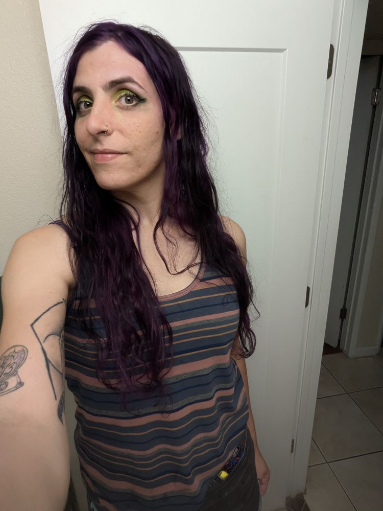 Selfie of Rayna in front of a white hallway door, you can see her long brown and purple hair and rust/blue striped tank top. Her eyeshadow is green "trash classic" themed in celebration of the new Frankie and the witch fingers album. This is after going to see Frankie and the witch fingers 
