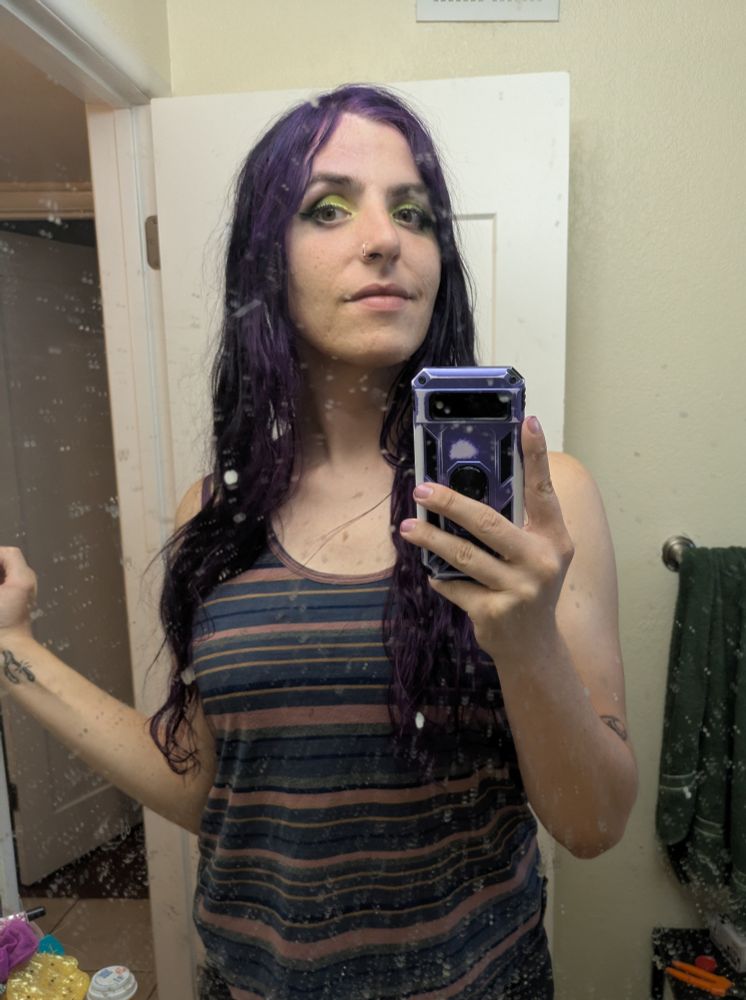 Rayna in front of her bathroom mirror, with green "trash classic" themed eyeshadow in celebration of the new Frankie and the witch fingers album. She has purple and brown hair and a striped rust/blue shirt. This is before going to see Frankie and the witch fingers 