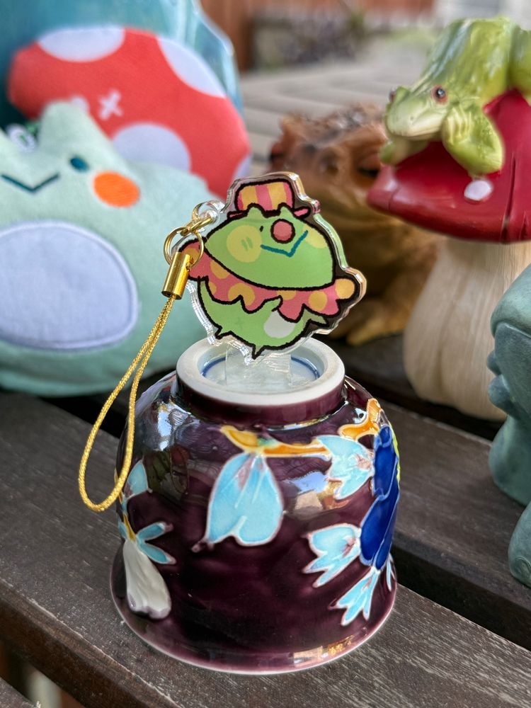 An acrylic phone charm of a clown standing on a cup stage surrounded by frogs