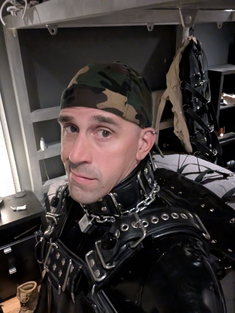 A gimp smirking with a backwards camo baseball cap, chonky collar, chonky chest harness, and latex catsuit 