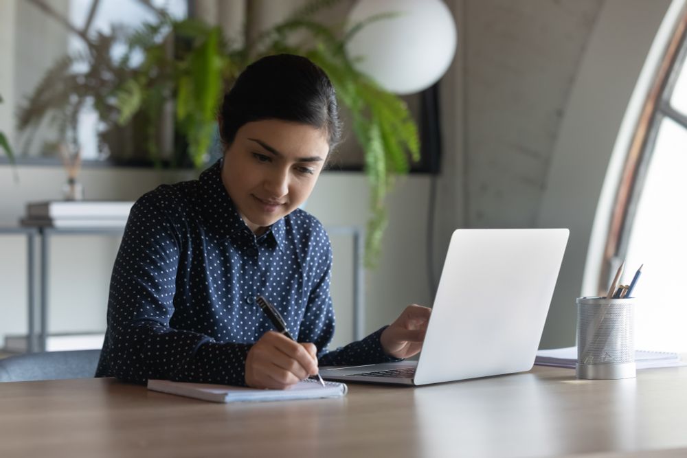 Woman employee taking notes on a notepad while on her laptop in a workspace.