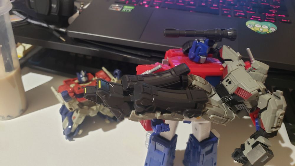 Optimus Prime Comander Class figure (g1 version) holding a Megatron Deluxe Class figure (transformers one version) while an Optimus Prime Deluxe Class figure (transformers one version) is on its knees screaming to the air with its arms raised.