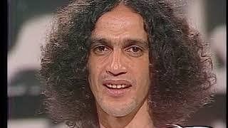 Imagem de um homem pardo com cabelo castanho cheio e ondulado na altura do queixo. Trata-se de um trecho de uma entrevista do Caetano Veloso em que ele está prestes a perder a paciência com a burrice dos interlocutores. 