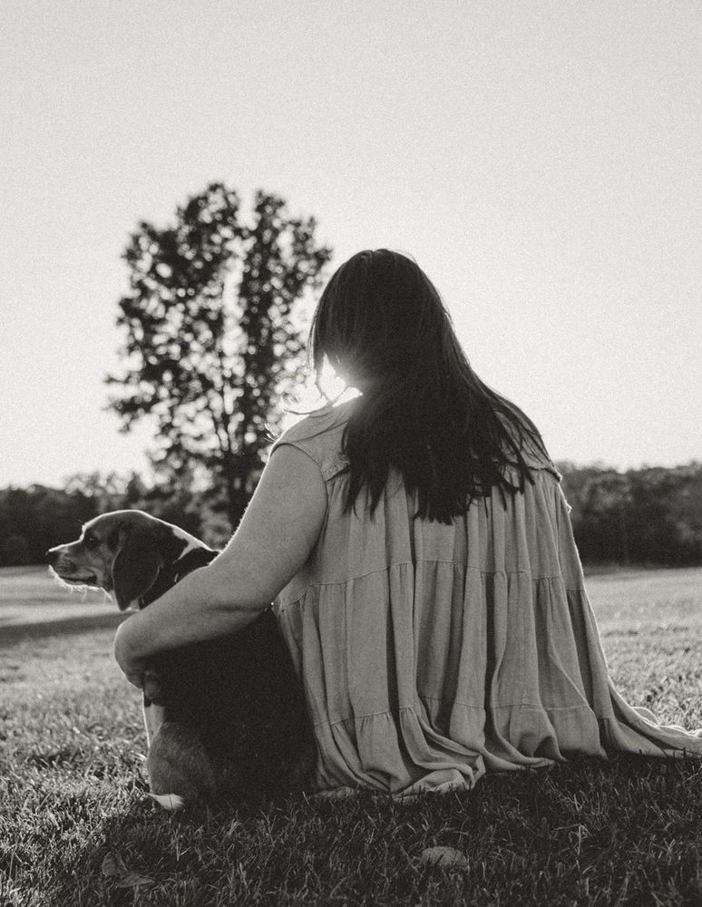 Gray tone photo of a woman with long dark hair sitting on the grass next to her beagle service dog with her arm around him. He is looking towards the left with a smirk on his face. She is looking away from the camera towards the sunset and a tree. 