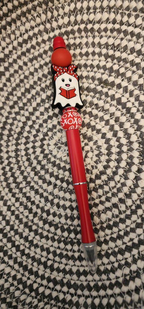 Red pencil with solid red and red with white "xoxo" round silicone beads next red rhinestone bead and with a white ghost silicone focal bead holding a red book and wearing a red with white polka dot bandana; sitting in a gray and white basket background.  