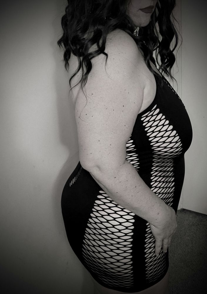Black and white photo. Side view of Poly in her fishnet dress. Its tightness is only eclipsed by its shortness. If only she could wear this thing out of the house. Alas... There are public indecency laws still.