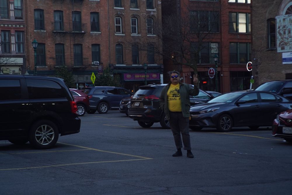 A wide shot of a man standing in a parking lot with cars in it. The city block buildings are in the background, there are no leaves on the trees, but the city is filled with life. The man is also holding up a peace sign.
