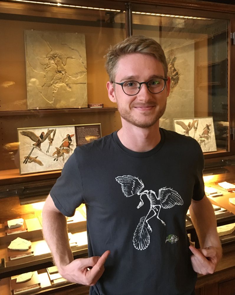 A young man standing in front of a museum showcase with an archaeopteryx fossil in a stone slab. He's pointing at his t-shirt which shows a drawing of the same fossil.