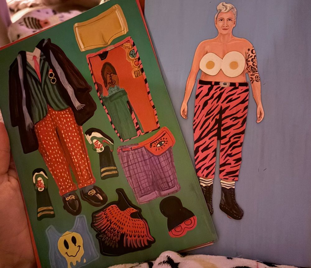 Sheets of magnetic outfits and little magnetic people. Off to the right a blonde magnet person with boobs is wearing combat boots, loud hot pink and black striped pants, and a sunny side up egg bra