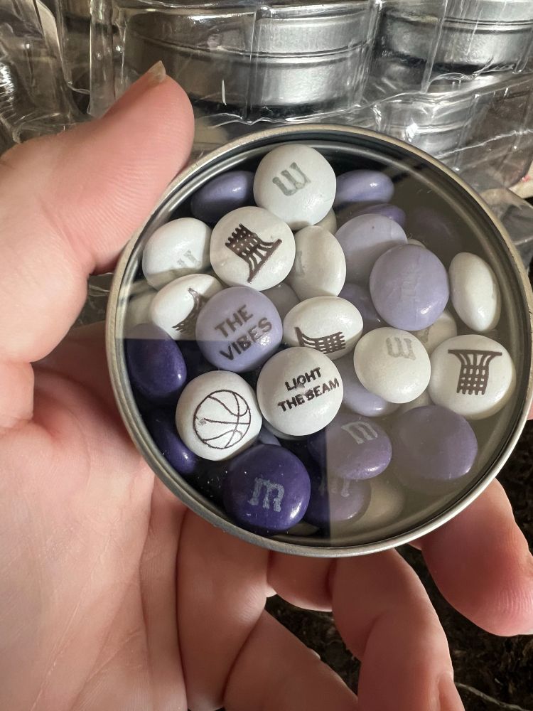 A hand holding a tin of m&m candies in purple, light purple and white. The candies are decorated with basketball icons and the phrases “light the beam” and “the vibes”. 
