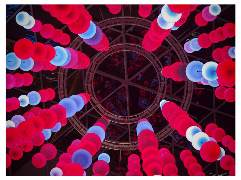 Colour photo looking up at a large round sky light, with red, blue and white illuminated orbs hanging down.