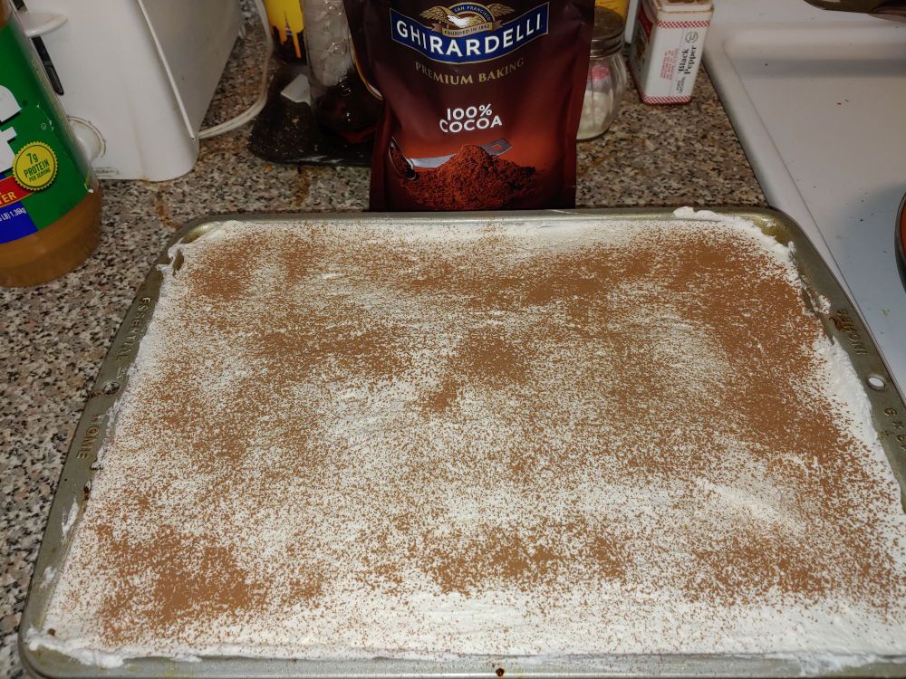 Completed tres leches cake in pan soaking in milk, covered with whipped cream and sprinkled with cocoa powder.