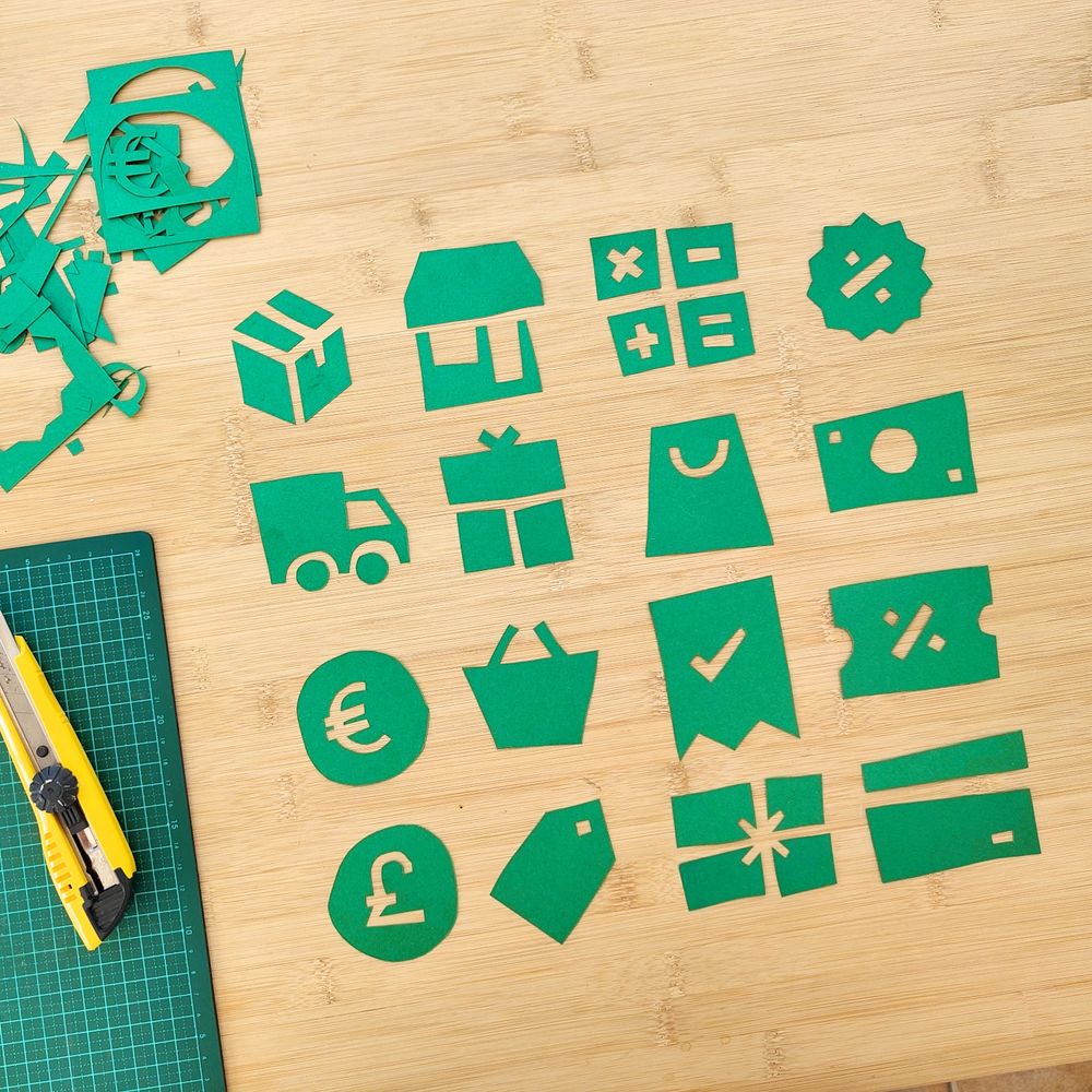 16 playful cutouts of e-commerce related icons, placed in a grid of 4 by 4 on a bamboo tabletop.