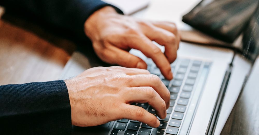 Close-up of a person typing on a laptop at a wooden desk, with documents, a pen, and a smartphone nearby.