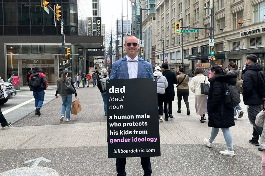 Person holding a sandwich-board sign with a faux dictionary definition that reads "Dad. noun. a human male who protects his kids from gender ideology."