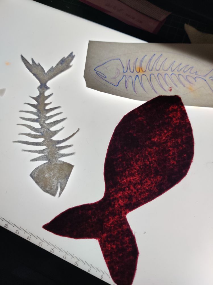 Photo de deux découpes de feutrine bordeaux et blanche sur une table lumineuse allumée. Il y a aussi un petit croquis du projet posé à côté.
La partie rouge représente la silhouette d'un poisson sans détail, et la partie blanche le squelette du poisson avec toutes les arrêtes.