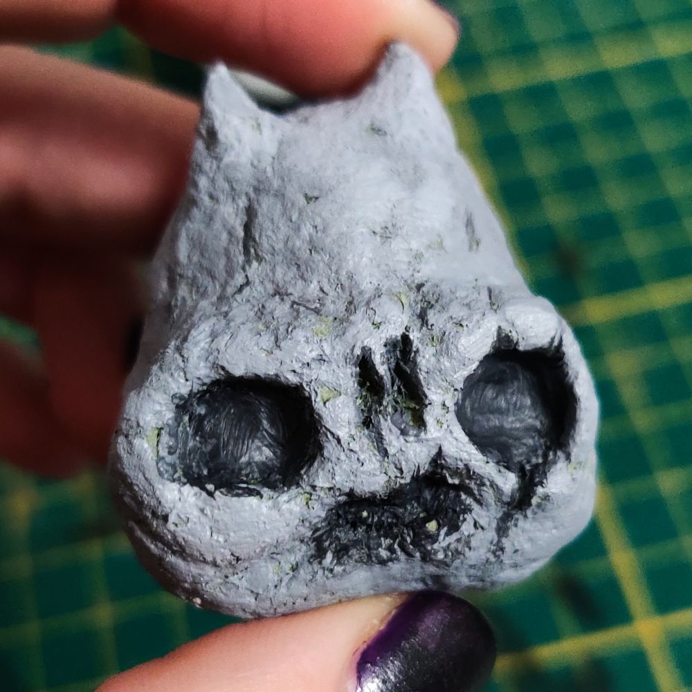 Photo d'une sculpture d'un tout petit crâne avec deux petites cornes. Il est gris avec quelques reliefs plus clairs ou plus foncés. Il a pas encore de dents devant.