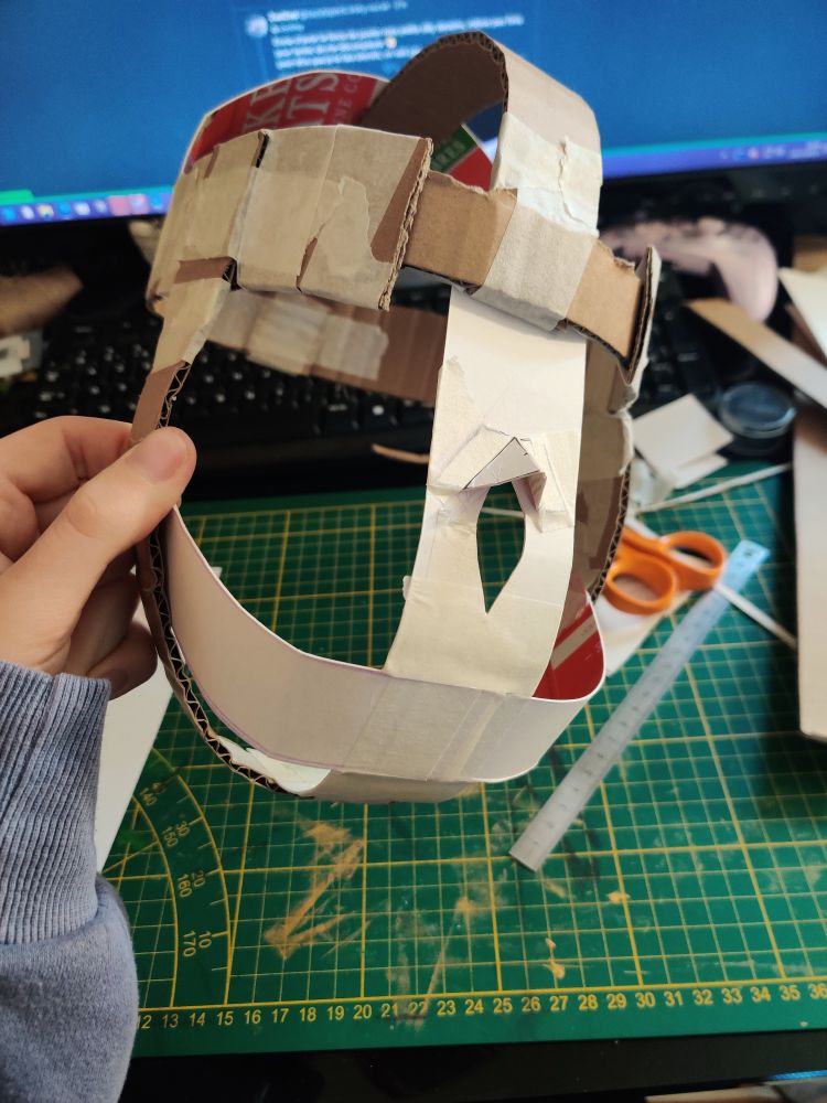Photo d'une construction rudimentaire en carton d'une base de masque. Certaines bandes de carton s'entrecroisent afin d'englober la circonférence du crâne, jusqu'au dessous de la mâchoire. Il est presque entièrement de face, un peu de trois quarts. Tout est tenu par du scotch de peintre, mis à part le relief d'une arrêté du nez qui est aussi collé pour plus de maintient.