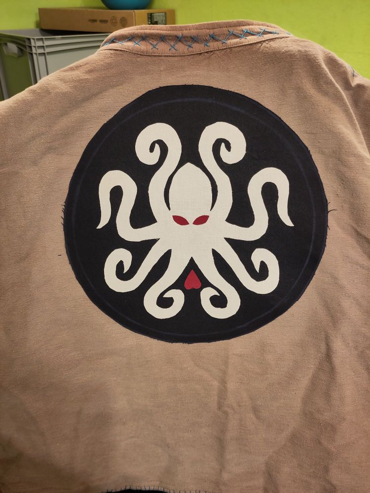 Der jolly der Kraken (weißer Tintenfisch mit roten Augen und rotem Herz darunter auf dunkelblauem Grund) auf einer ausgeblichen braunen weste 