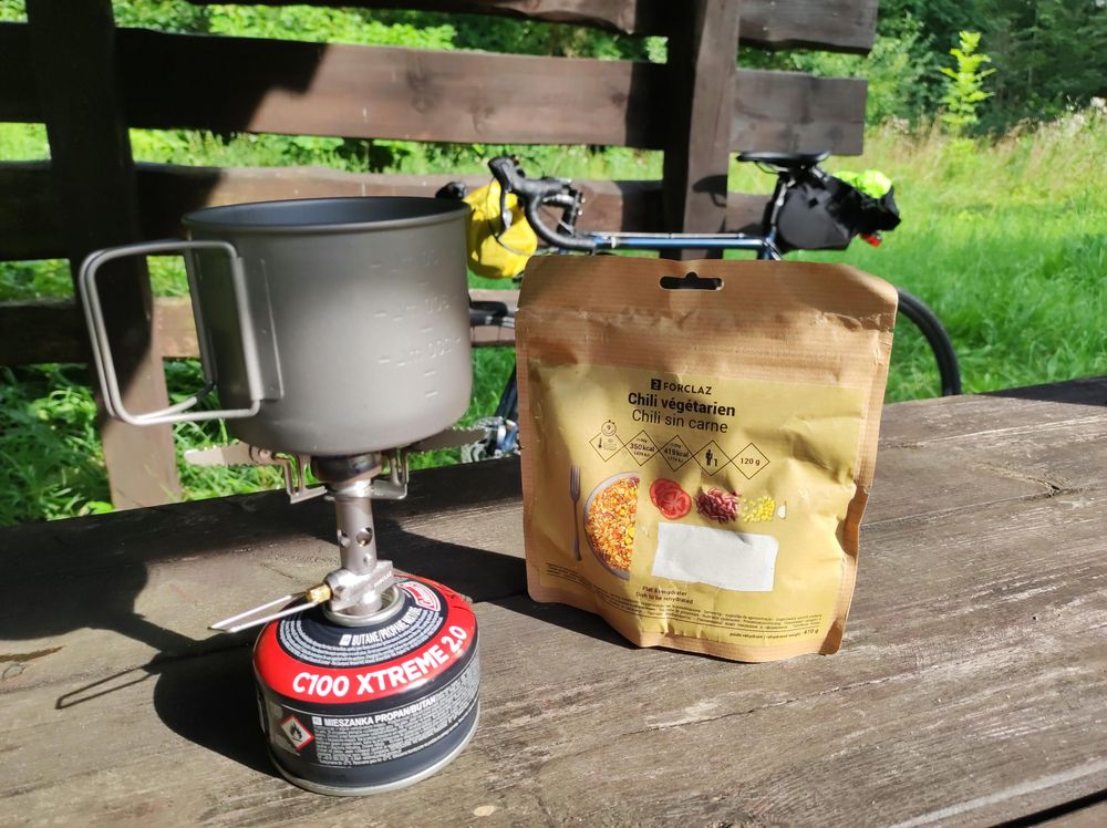 Na drewnianym stole pod wiatą stoi mała kuchenka gazowa, a na niej kubek, w której gotuje się woda. Obok jest paczka wegetariańskiego chili sin carne.
