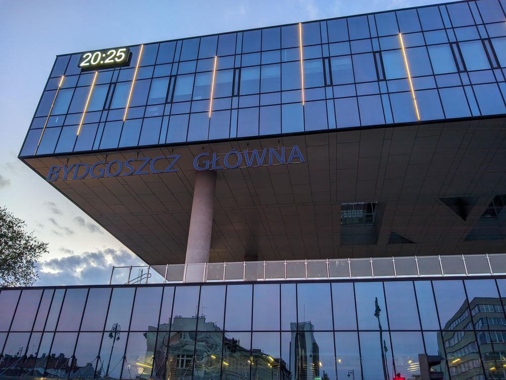 Budynek dworca kolejowego Bydgoszcz Główna, składający się z dwóch prostopadłościanów o przeszklonych bokach. W dolnym odbija się wieczorne miasto, góry jest ciemnoniebieski od odbicia nieba. W lewym górnym rogu znajduje się zegar pokazujący 20:25.