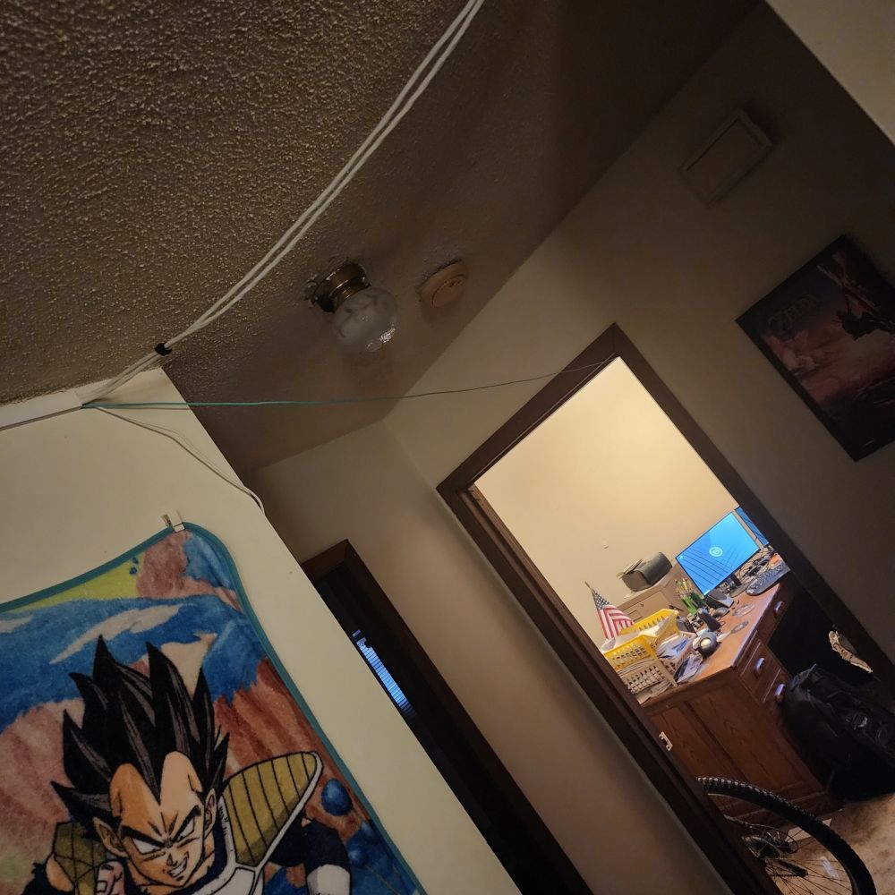 A poorly-lot picture showing the blue Multimpde fiber exiting the corner of some conduit on the ceiling.

There are a few white speaker cables velcroed together going across this gap towards the camera for the rear surround sound speakers. The blue fiber optic cable hangs mostly freely, lowering slightly to go into the top of the doorframe of the office. There is a rear bike tire partly inside the office doorway, and further until the office is a desk with one of two monitors showing the default Linux Mint desktop background.