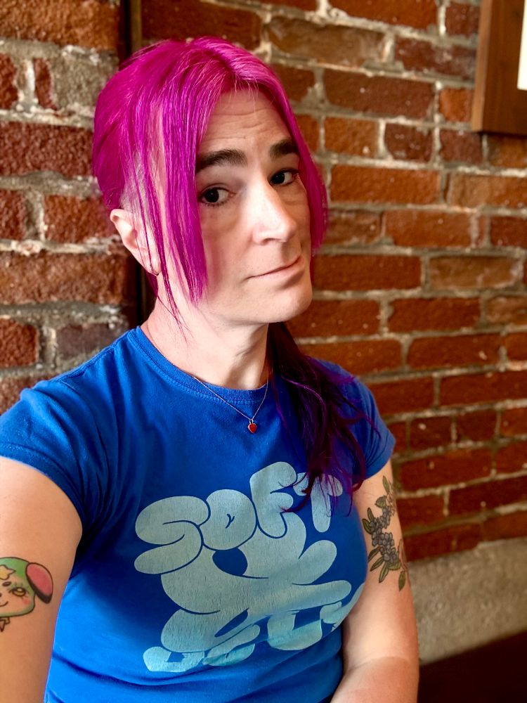 becs sitting in front of a brick wall, in a blue tee with pink hair, & slightly smiling