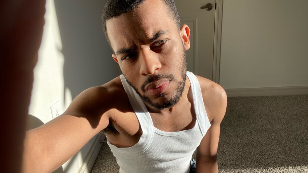 Aaron in a white tank top and blue jeans squatting on the floor and holding the camera lens.