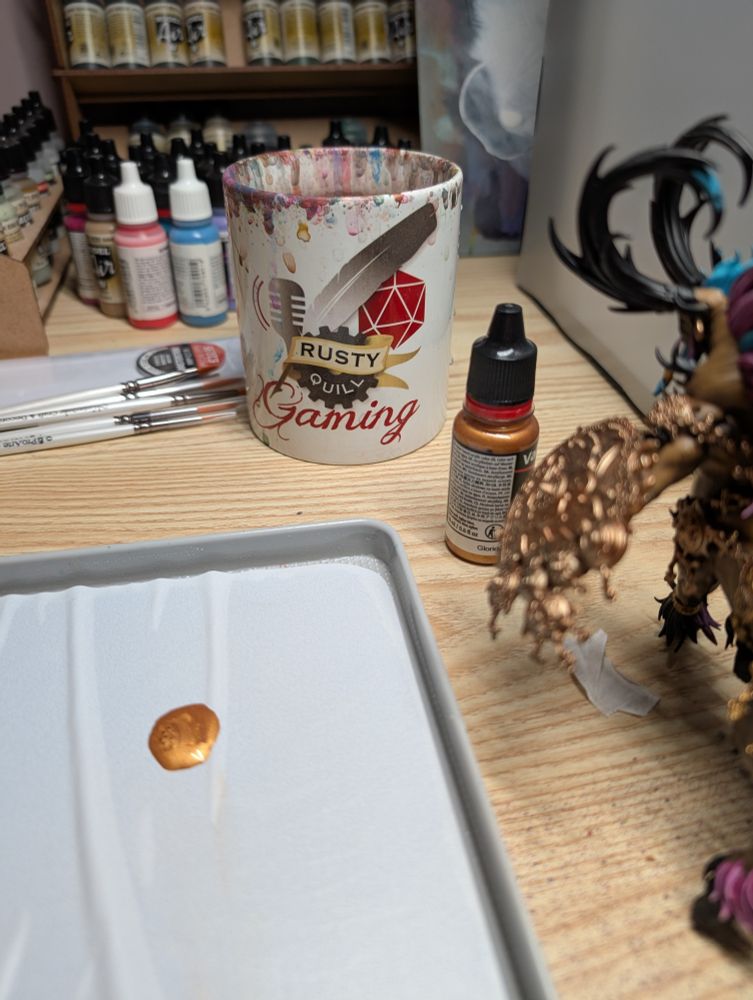Photo of a desk set up for painting miniatures. The focus of the image is an old Rusty Quill Gaming mug which is being used to hold water for paint brushes. The mug is white and stained with many different colours of paint.