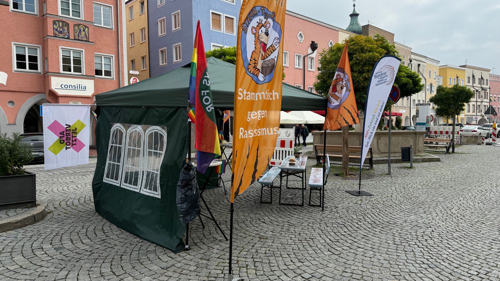 Auf dem Bild sieht man einen grünen Pavillon auf dem Stadtplatz von Mühldorf, an dem Pavillon sind Fahrräder befestigt. Davor steht eine Bierbank in hellblau. Rund um den grünen Pavillon sieht man Fahnen vom Stammtisch gegen Rassismus, Altöttinger Bündnis für Respekt, Vielfalt und Demokratie und von Fridays for Future. 