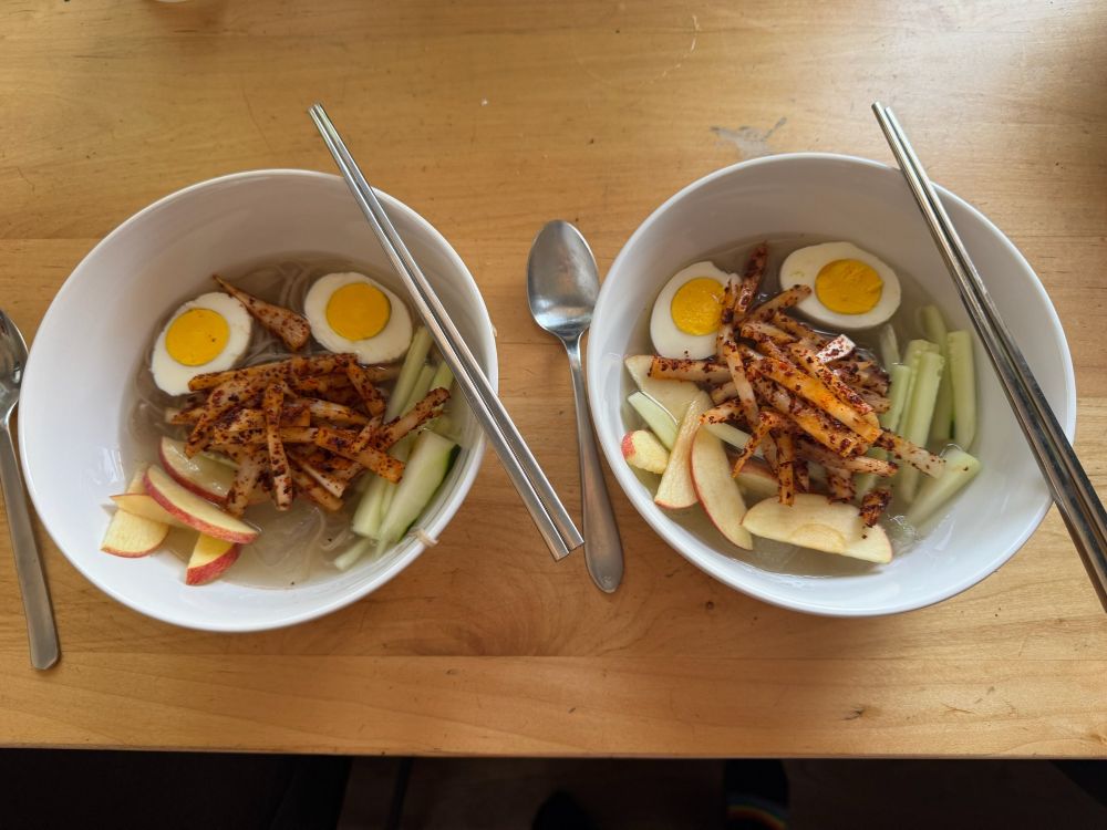 김치말이국수 Kimchimari-guksu. 
Two white bowls showing 2 halves of a hard boiled egg, sliced apples and cucumbers, with seasoned kimchi radish in the centre. Icy chunks of soup can be seen. 
A pair of chopsticks and a spoon with each bowl. 