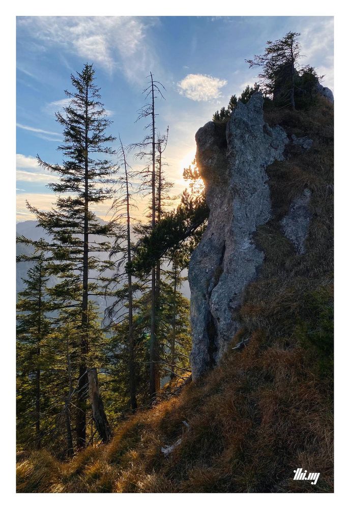 The silhouette of a standalone rock needle and some nearby trees along the ridge. The sun and some mountains are partially visible from behind the rock. Beautiful red/brown grasses.