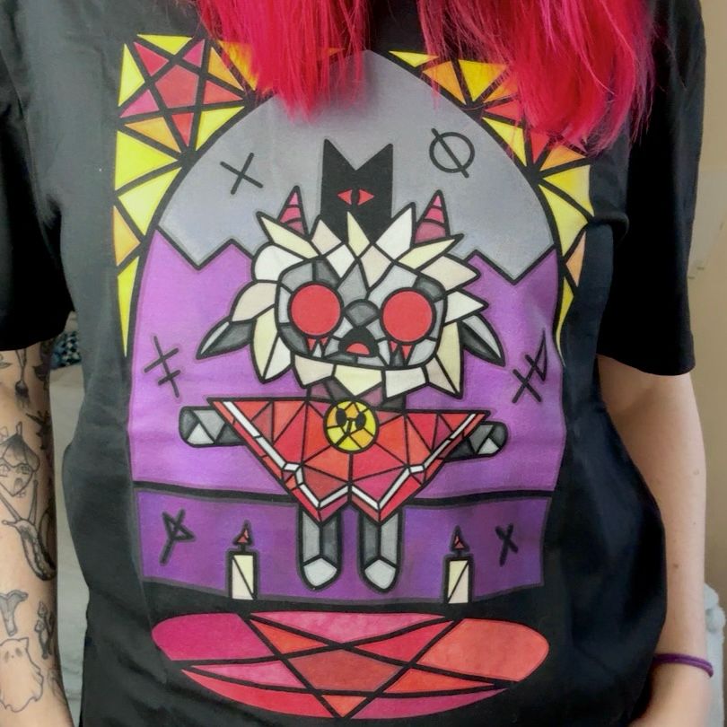 A woman modeling a stained glass style cult of the lamb tee. The artwork shows the lamb video game character floating above a pentagram. The T-shirt is crew neck, black, and relaxed fit. The image is cropped at the models head and waist. Her dyed pink hair is showing at the top. Her left arm has patchwork tattoos and her right has a hair scrunchie.