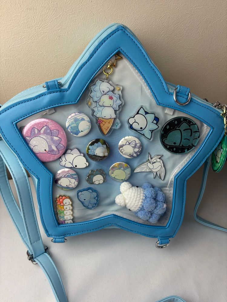 A star shaped cross body ita bag filed with snom art. There are button badges, enamel pins, acrylic keychains, acrylic pins and a crochet Snom plushie.